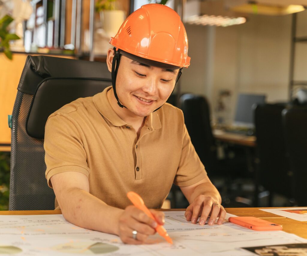 pexels-photo-8278900-8278900 Smiling engineer in hard hat studies blueprints at an office desk, highlighting construction and planning.